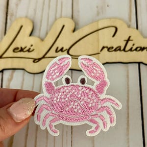 May include: Embroidered pink crab patch with white trim. The crab has detailed claws and body, with brown eyes. The patch is held in front of a wooden sign that reads "Lexi Lu Creations".