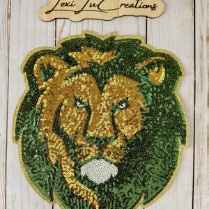 May include: A sequined lion head patch in gold and green. The lion's mane and face are detailed with gold sequins, while the background is green. A wooden sign with the text "Lexi Lu Creations" is above the patch.