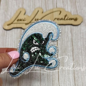 May include: A sequined patch shaped like a stylized wave with a dark green and black creature face featuring an angry expression. The design includes white and light blue sequin accents, a silver border, and a white embroidered outline.