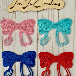 May include: Four iron-on patches in the shape of a bow with a stethoscope. The bows are pink, turquoise, blue, and red. The stethoscopes are silver.