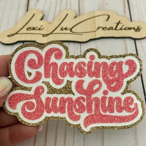 May include: A pink and white glitter sticker with the words "Chasing Sunshine" and a gold glitter border. The sticker is held in a hand, with a wooden sign in the background that says "Lexi Lu Creations".
