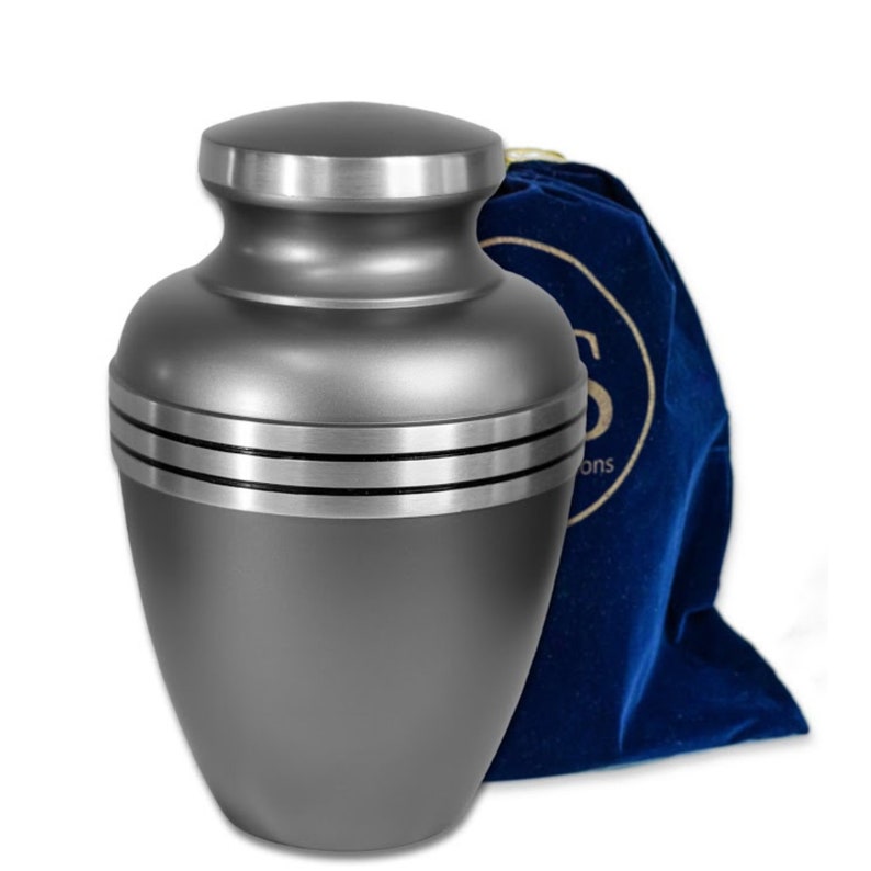 May include: A gray metal cremation urn with a silver trim and three silver bands around the middle. The urn comes with a blue velvet bag.