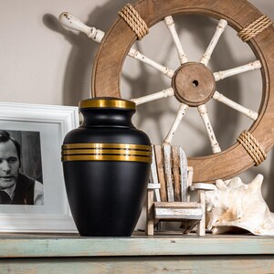 May include: Black urn with gold trim, sitting on a wooden shelf with a nautical theme. The urn is decorated with three gold bands around the middle.