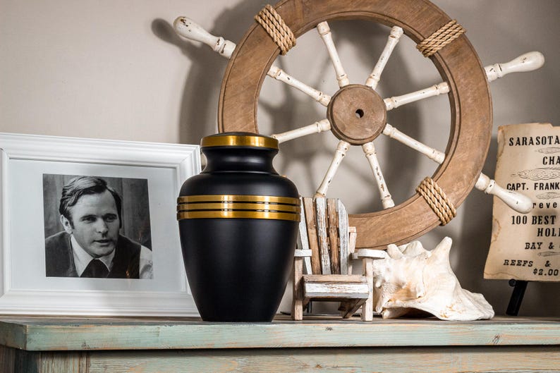 May include: Black urn with gold trim, sitting on a wooden shelf with a nautical theme. The urn is decorated with three gold bands around the middle.