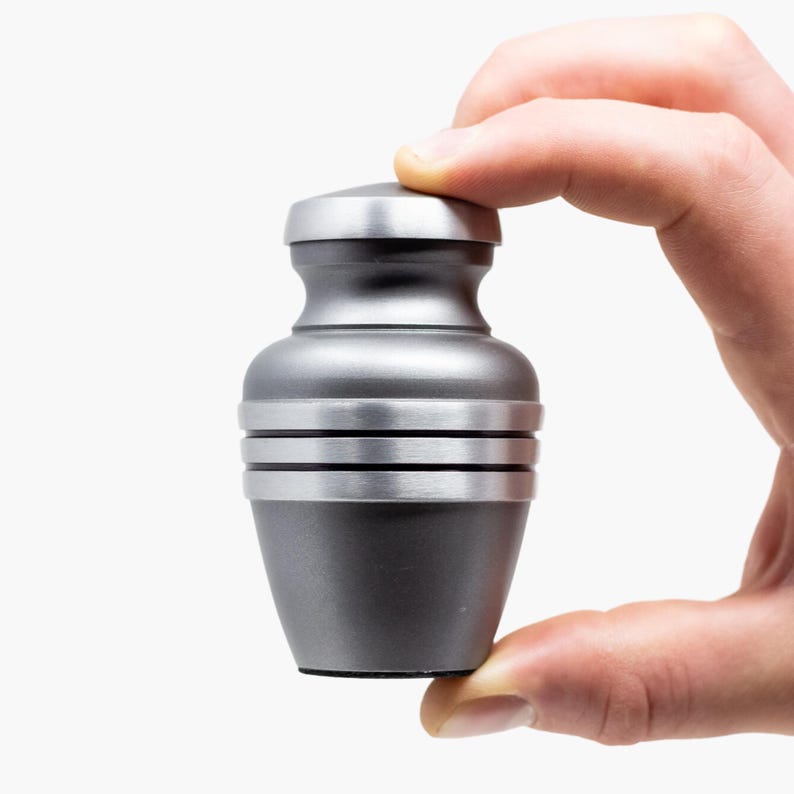 May include: A silver metal urn with a smooth, rounded top and a textured, ridged body. The urn is held in a hand.
