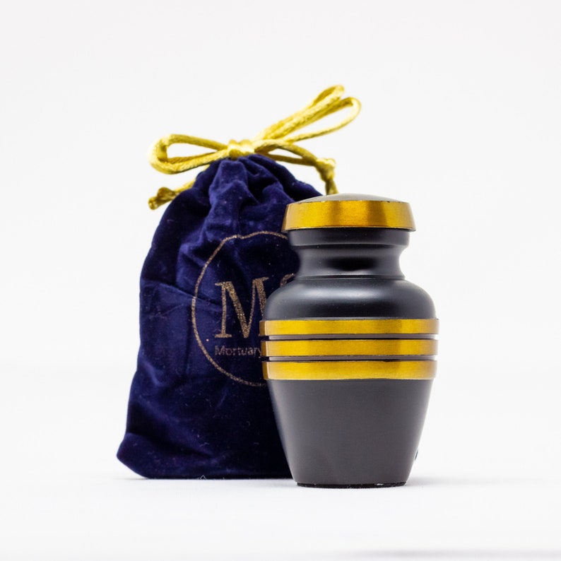 May include: Black cremation urn with gold trim and a velvet carrying bag. The bag has a gold drawstring and a logo that says "Mortuary Supplies".