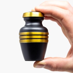 May include: A small black urn with gold trim. The urn is being held in a hand.