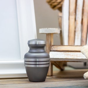 May include: A small, gray metal urn with silver bands. The urn is sitting on a wooden surface.