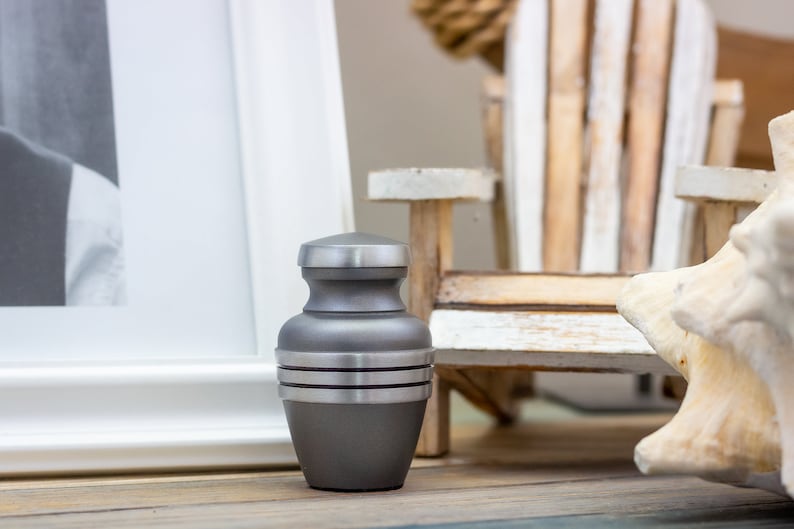 May include: A small, gray metal urn with silver bands. The urn is sitting on a wooden surface.