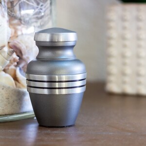 May include: A silver metal urn with a smooth, rounded body and a flat lid. The urn has three silver bands around the middle.
