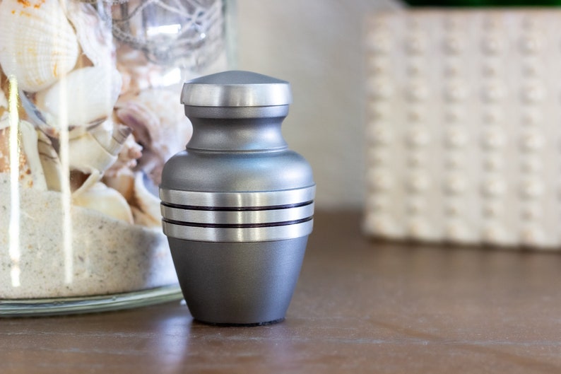 May include: A silver metal urn with a smooth, rounded body and a flat lid. The urn has three silver bands around the middle.