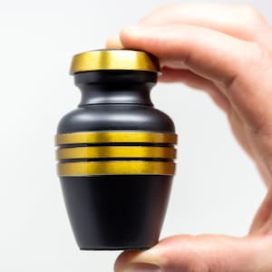 May include: A small black urn with gold trim. The urn is being held in a hand.