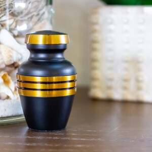 May include: A small black urn with gold trim. The urn is sitting on a wooden table.