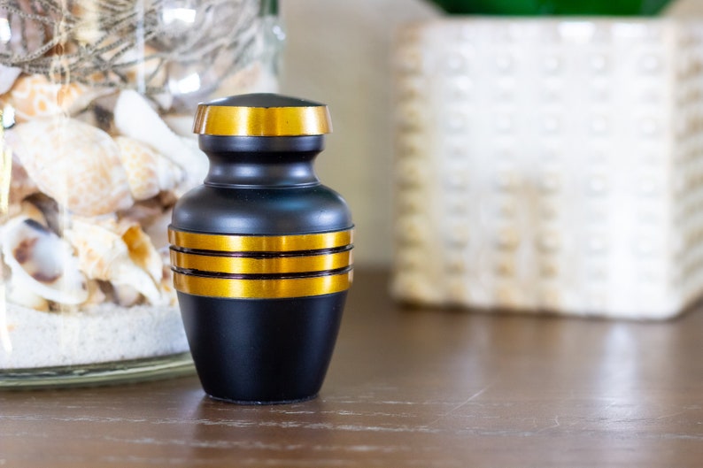 May include: A small black urn with gold trim. The urn is sitting on a wooden table.