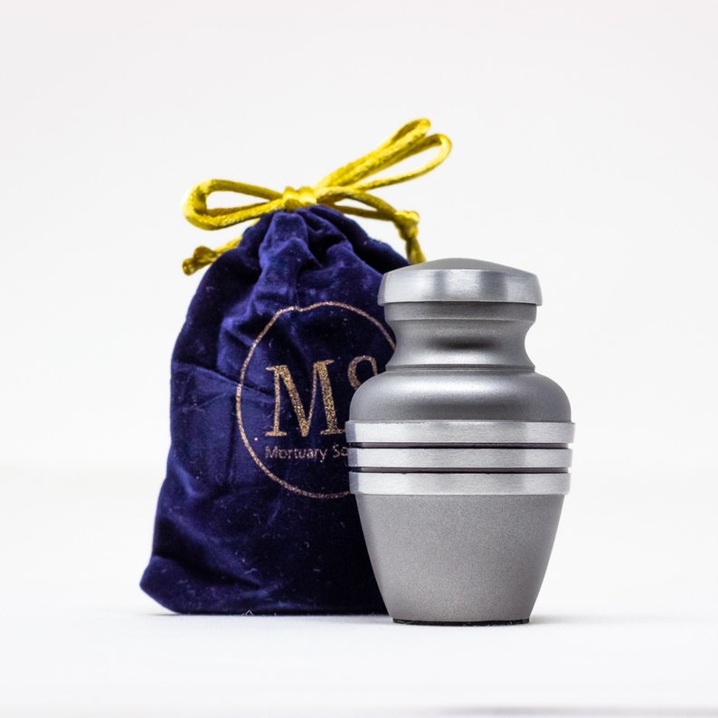 May include: A silver metal cremation urn with a satin finish and three silver bands around the middle. The urn is sitting on a blue velvet bag with a gold drawstring.