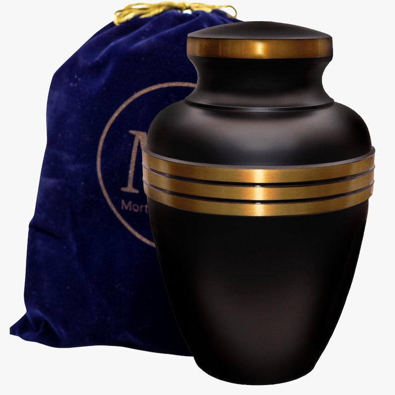 May include: Black metal urn with gold trim and a blue velvet carrying bag. The bag has a circular logo with the word "Mort" on it.