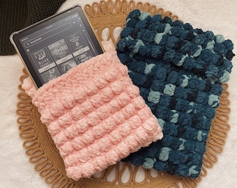 May include: Two crocheted pouches in pink and teal with a textured bubble design. The pouches are on a woven straw mat and contain an e-reader.