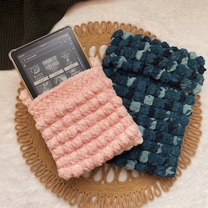 May include: Two crocheted pouches in pink and teal with a textured bubble design. The pouches are on a woven straw mat and contain an e-reader.
