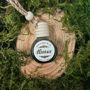 Könnte beinhalten: Ein Auto-Lufterfrischer mit dem Text "Alistair" auf einem weißen Etikett, umgeben von einem schwarzen Rand. Die Flasche ist klar mit einem Holzdeckel und einer Schnur zum Aufhängen. Der Hintergrund zeigt grünes Moos und eine Holzscheibe.