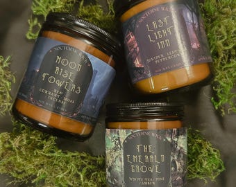 May include: Three amber glass soy wax candles with black lids and labels. Labels read Nocturne Wick Moon Rise Towers Red Currant/Moss/Incense, Last Light Inn Juniper/Leather/Peppercorn, and The Emerald Grove White Tea/Pine/Amber. Each candle is 200g.