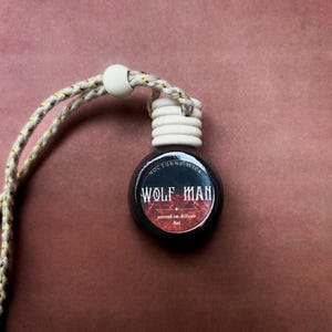 May include: A small, round, black car air freshener with the text "WOLF MAN" in a gothic font. The freshener has a white wooden cap and a braided cord with gold accents. The background is a solid, muted reddish-brown.
