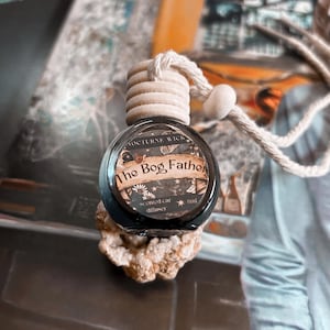 Puede incluir: Un ambientador para coche con tapa de madera y botella de cristal transparente. La etiqueta dice "The Bog Father" con el texto "Nocturne Wick" encima. El ambientador está suspendido por una cuerda blanca.
