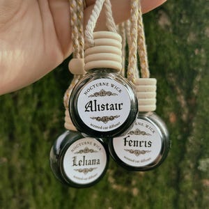 May include: Three small, round, black car air fresheners with white labels featuring names and the text "Nocturne Wick scented car diffuser." Each bottle has a wooden cap and is suspended by a beige cord with gold accents. The names on the labels are Alistair, Leliana, and Fenris.