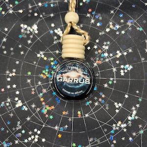 Peut inclure: Un diffuseur de parfum pour voiture avec une étiquette noire et bleue portant l'inscription "GARRUS" et "scented car diffuser". Il est suspendu à une ficelle beige et une perle en bois. Le fond est une surface sombre avec un motif de constellation et des confettis en forme d'étoiles.