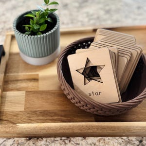May include: A set of wooden shape puzzle cards with a star cutout and the word "star" printed on the card. The cards are in a brown wicker basket on a wooden tray.