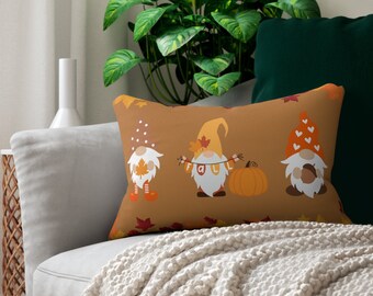 Puede incluir: Una almohada de sofá marrón con un tema otoñal con tres gnomos sosteniendo una pancarta que dice "Fall". Los gnomos llevan sombreros rojos y blancos y tienen barbas blancas. La almohada está decorada con hojas naranjas y una calabaza.
