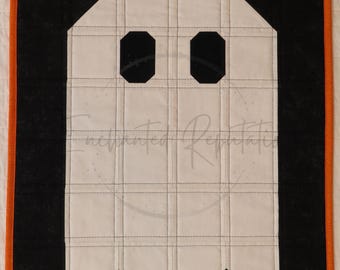Puede incluir: Un edredón de pared rectangular que presenta una forma de fantasma blanco centrada sobre un fondo negro. El fantasma tiene dos ojos negros octogonales y está enmarcado por un borde naranja fino. La tela tiene un patrón de cuadrícula.