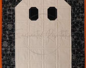 Puede incluir: Un tapiz acolchado rectangular que presenta una forma de fantasma blanca central con dos ojos octogonales negros. El fantasma se sitúa sobre un fondo negro con estampado de telarañas blancas, enmarcado por un borde de tela naranja.