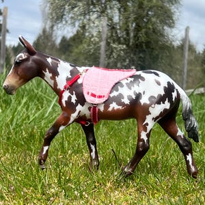 Puede incluir: Figura detallada de una mula con pelaje manchado marrón y blanco, que lleva una montura rosa y un arnés rojo. La mula posa en un campo de hierba, con un fondo borroso de árboles y un cielo nublado.