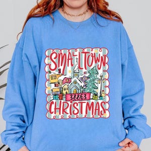 May include: A light blue crewneck sweatshirt featuring a graphic of a snowy village with houses, a church, a Christmas tree, and a streetlamp. The text reads "SMALL TOWN" and "CHRISTMAS" in red, with "38863" on a pink rectangular sign.