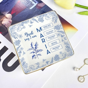 Puede incluir: Un plato cuadrado de cerámica blanca con un diseño floral y de mariposas azul. El plato tiene un borde dorado y las palabras "God say I am MARIA" en azul. El plato también tiene las palabras "MADE NEW", "ACCEPTED", "RESTORED", "INVITED" y "ABUNDANT".