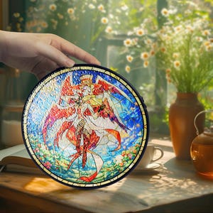 May include: A round, stained-glass art piece featuring a colorful angel design with red, white, and blue hues. The artwork is held by a hand, showcasing the intricate details and vibrant colors. The piece has a black border and is likely a decorative item.