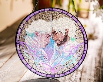 May include: Round stained glass window art featuring two figures with white hair and beards, in a tender embrace. The artwork uses pastel colors, including purple, blue, and green, with a black border and chain for hanging. The glass has a textured appearance.