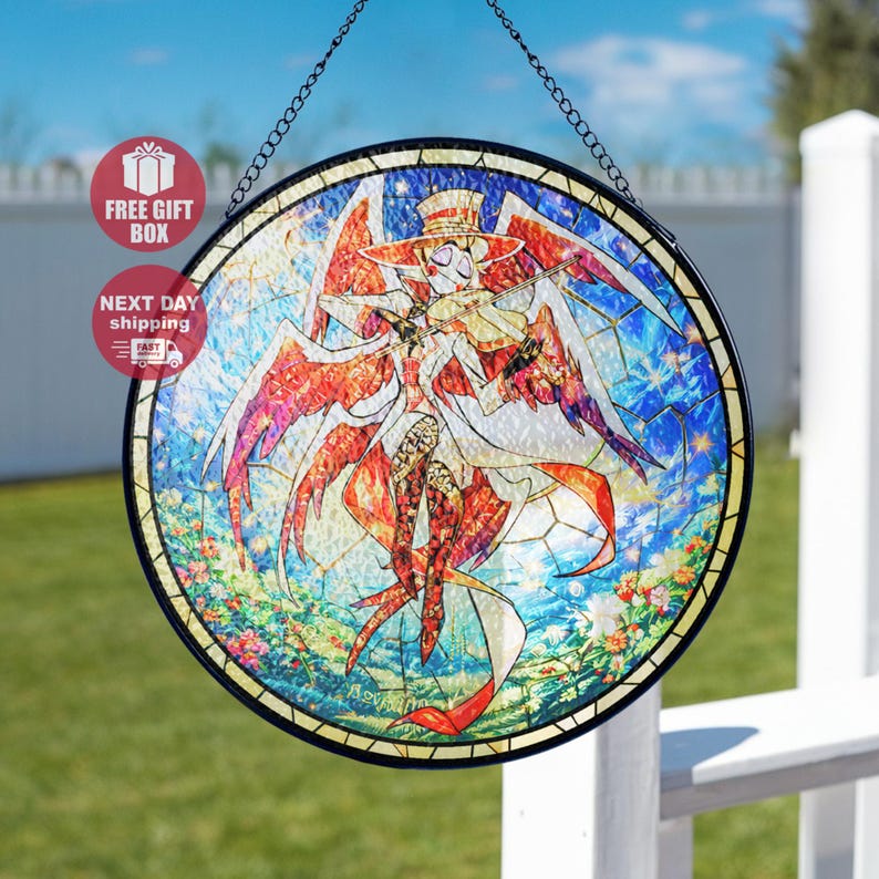May include: A round stained glass window featuring a character playing a violin, with a hat and wings. The artwork uses a vibrant palette of red, orange, blue, and white, with a floral border. The window is framed in black and hangs from a chain.
