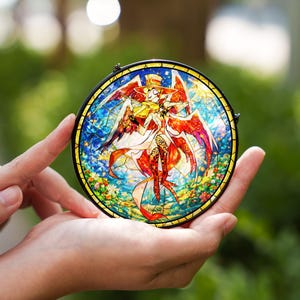May include: A round stained glass art piece featuring a colorful angel-like figure with wings, a hat, and a violin. The artwork is set against a blue and yellow background with a black border. The piece is held in open hands.