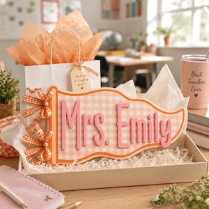 May include: A pennant-shaped wooden sign featuring the name Mrs. Emily in raised pink lettering on a light orange gingham background. The sign is decorated with two orange and white checkered bows and is presented in a gift box with tissue paper.