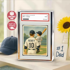 Puede incluir: Una obra de arte enmarcada con temática de béisbol que muestra a un padre y su hijo en un campo de béisbol. La camiseta del padre dice "HENRY 10", y la del hijo "WILSON 07". La obra está encerrada en un bloque acrílico transparente, con el texto "2026 FAMILY LEAGUE NO. 1 DAD".