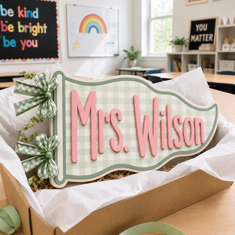 May include: A pennant-shaped wooden sign featuring a light green and white gingham pattern with the text Mrs. Wilson in raised pink lettering. The sign is decorated with two green and white gingham bows and green ribbon curls on the left side.