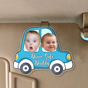 Könnte beinhalten: Ein blauer, autoförmiger Sonnenblendenclip mit zwei Babygesichtern in den Fenstern. Das Auto hat gelbe Räder und den Text "Drive Safe Daddy" auf der Vorderseite.