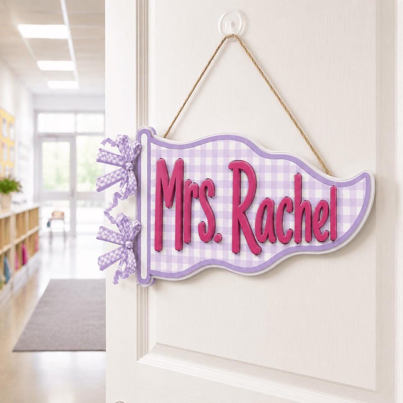 May include: A pennant-shaped door sign featuring a light purple and white gingham pattern. The sign displays the text "Mrs. Rachel" in raised, dark pink lettering. Two gingham bows are attached to the left side, and a jute twine hanger is included.