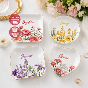 Könnte beinhalten: Vier weiße Keramikschalen mit goldfarbenen Rändern, jede mit einer individuellen Blumenillustration und einem personalisierten Namen versehen. Die Schalen sind quadratisch, rund und herzförmig und mit rosa, gelben und violetten Blumen verziert.