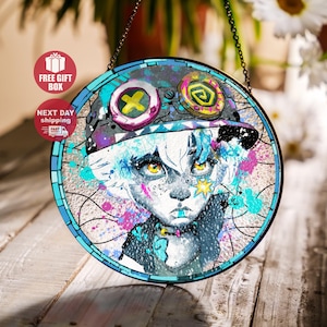Puede incluir: Obra de arte circular en vidriera que representa un personaje de estilo anime con ojos amarillos y una estrella. El diseño incorpora un fondo abstracto vibrante con salpicaduras azules, rosas y moradas. La pieza está enmarcada con un borde azul y suspendida por una cadena.