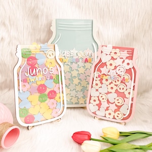 以下が含まれることがあります： 透明なプラスチック製のインサートが付いた、木製のメイソンジャー型の報酬チャートが3つあります。ジャーは、カラフルな星、ハート、スマイリーフェイスで飾られています。ジャーには、「Juno's Reward Jar」、「Classroom Reward Jar」、「Juno's Reward Jar」とラベルが貼られています。