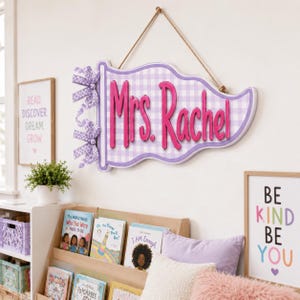 May include: A wall-hanging pennant sign featuring a light purple and white gingham pattern with the text Mrs. Rachel in raised, bright pink lettering. The sign is suspended by a tan rope and decorated with two purple and white gingham bows.
