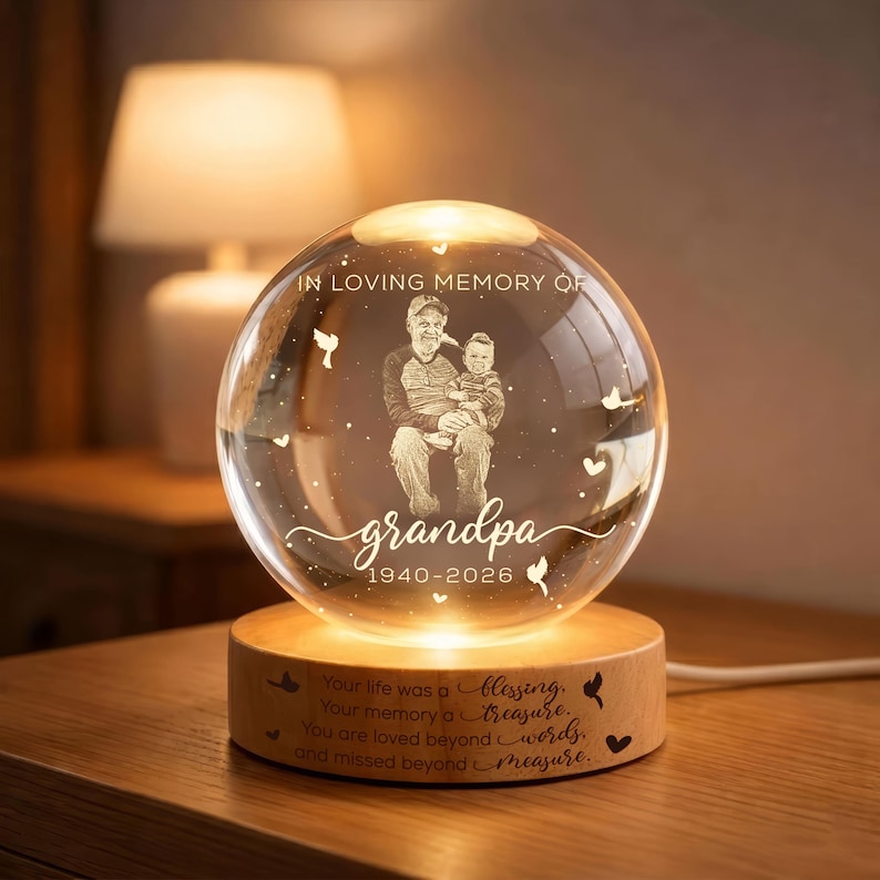 May include: A clear glass sphere with an engraved image of a man and child, lit from within. The sphere sits on a wooden base with engraved text: "IN LOVING MEMORY OF grandpa 1940-2026." Additional text on the base reads: "Your life was a blessing..."