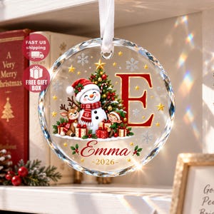 Könnte beinhalten: Ein kreisförmiger Weihnachtsbaumschmuck aus Glas mit einem Schneemann, einem Rentier und einem Elfen mit Geschenken. Das Design zeigt einen großen roten Buchstaben E, den Namen Emma und das Jahr 2026. Es hängt an einem weißen Band und ist mit goldenen Sternen verziert.